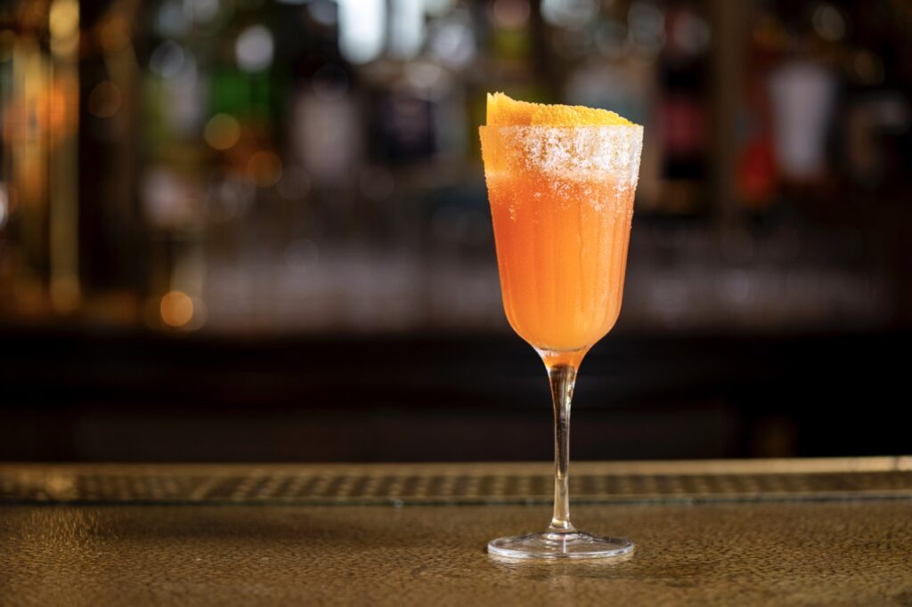 An orange cocktail sits at a bar with an orange rind in it.
