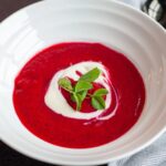 A bowl of vibrant red Polish Borscht beet soup, garnished with a swirl of crème fraîche and fresh pea shoots.