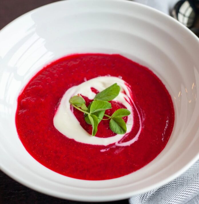 A bowl of vibrant red Polish Borscht beet soup, garnished with a swirl of crème fraîche and fresh pea shoots.
