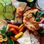 A bowl full of rotisserie buffalo chicken dip with pita bread, carrots, cucumbers, chips, and pretzels around it.