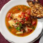 A white bowl filled with Baccalà fish stew with potatoes and tomatoes, served with a piece of grilled, charred bread on the rim.