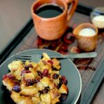 A plate of bread pudding with dried fruit and raisins, a fork resting on the plate, next to a mug of coffee and a small jug of cream on a dark wooden tray.