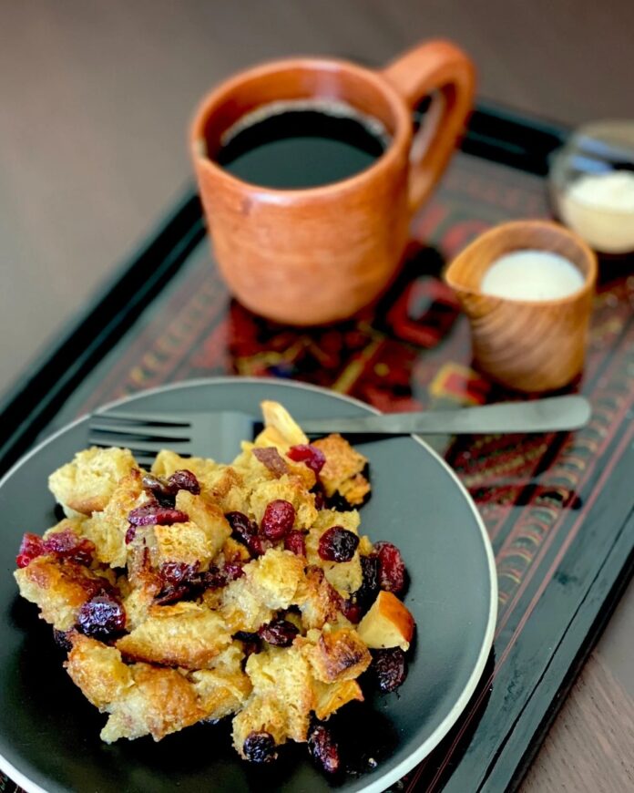 A plate of bread pudding with dried fruit and raisins, a fork resting on the plate, next to a mug of coffee and a small jug of cream on a dark wooden tray.