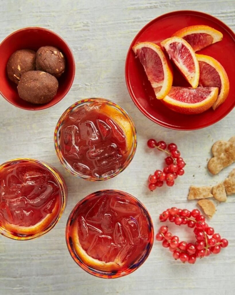 Three Orchard Negroni cocktails with blood orange wedges, garnished with red currants and surrounded by cookies.