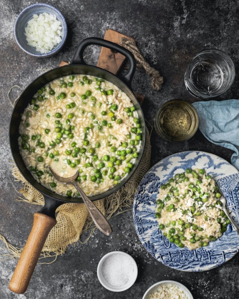 A creamy pot of Risi e Bisi (Venetian rice and peas) and a serving on a blue and white plate, garnished with Parmesan cheese and a side of chopped onion.