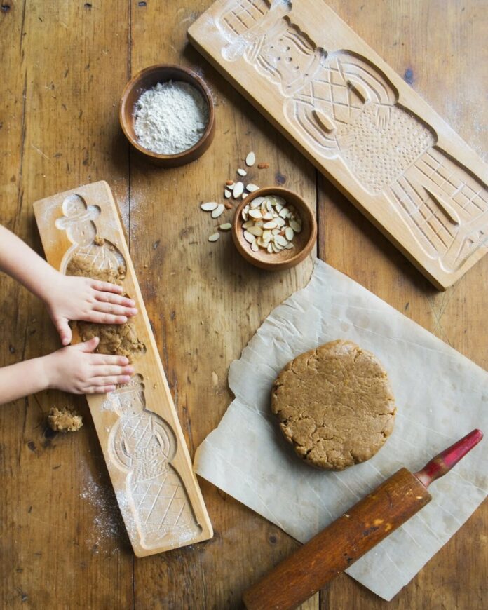 Two wooden gingerbread cookie molds, one of which a child's hands are pressing dough into, next to a ball of dough, a rolling pin, and small bowls of flour and sliced almonds.