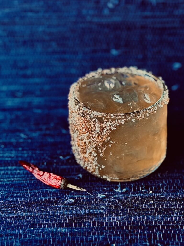 A rocks glass with The Boss rum cocktail in it with a salt and sugar rim. It sits on a blue surface beside a chile pepper.