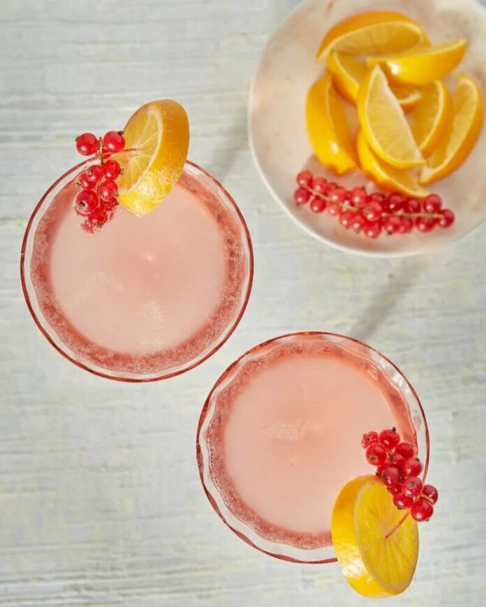 The Fitzgerald TABLE Magazine Two pink Fitzgerald gin cocktails garnished with red currants and lemon slices on a white wooden surface.