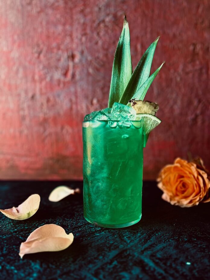 A vibrant aqua green cocktail in a tall glass with pineapple leaves, a pineapple wedge and lime wedge as garnish with an orange rose and rose petals in the foreground.