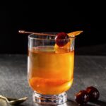 A short cocktail glass with the Witcher's White Honey using bourbon and orange peels on the inside. A stick with a luxardo cherry sits over top of the glass.