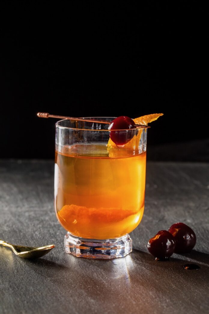 A short cocktail glass with the Witcher's White Honey using bourbon and orange peels on the inside. A stick with a luxardo cherry sits over top of the glass.