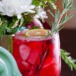 A red Yule Margarita cocktail on ice, garnished with a lime slice and a rosemary sprig, with holiday decor in the background.