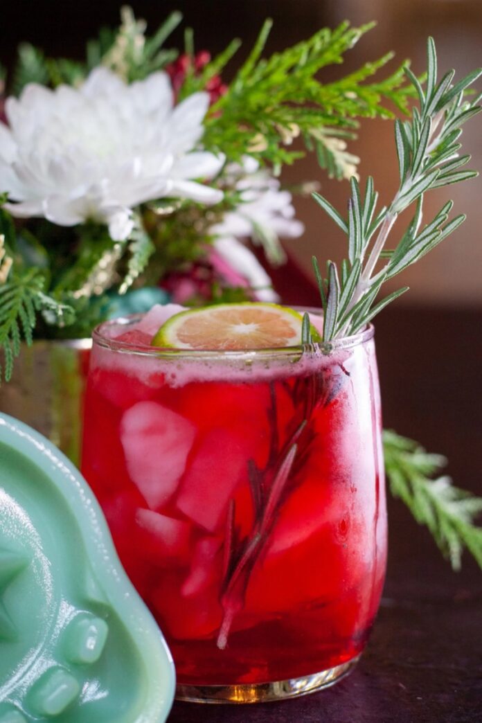 A red Yule Margarita cocktail on ice, garnished with a lime slice and a rosemary sprig, with holiday decor in the background.