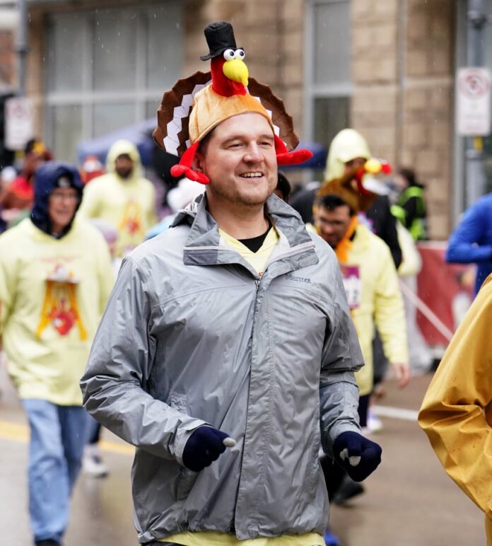 2025 Thanksgiving Events A man in a grey windbreaker jacket with a turkey hat running in a race.