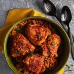 A bowl of Arroz con Pollo featuring rice and four bone-in chicken thighs topped with a rich red sofrito sauce and parsley, served with black metal spoons.