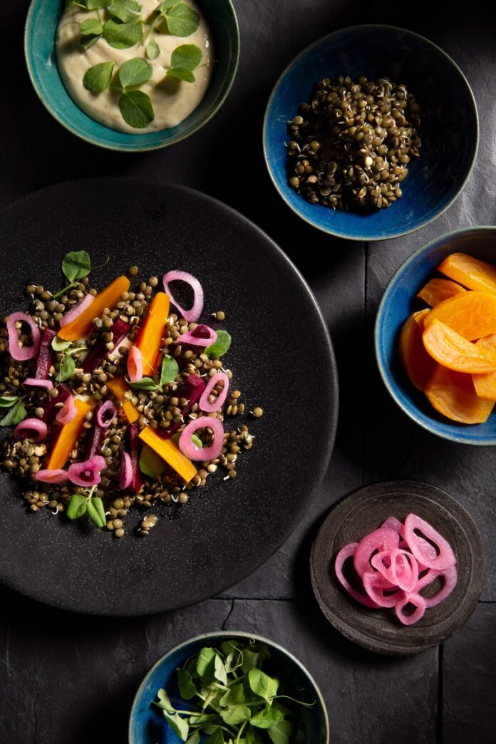 An overhead, dark food photography shot of a plate of beets, sprouted lentils, and tahini goat cheese custard, with small bowls of ingredients like pickled red onion and whole lentils surrounding it.
