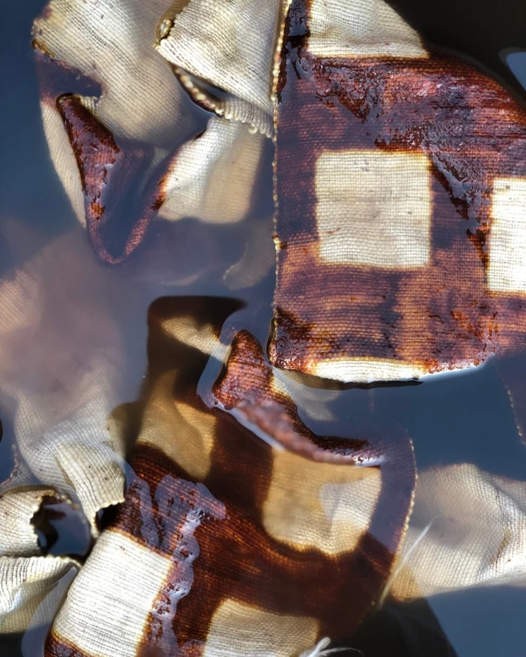 Woven cotton bògòlanfini mudcloth fabric soaking in a bath of dark liquid, showing brown geometric patterns bleeding into the white cloth during the careful cleaning stage of the dyeing process.