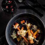 A gourmet salad with roasted carrots, chickpea fritters, and frisée lettuce, topped with vibrant red pomegranate seeds and harissa vinaigrette, served in a deep blue bowl with black flatware nearby.