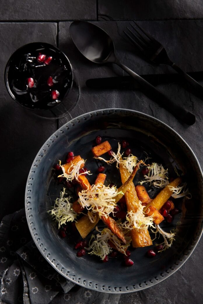 A gourmet salad with roasted carrots, chickpea fritters, and frisée lettuce, topped with vibrant red pomegranate seeds and harissa vinaigrette, served in a deep blue bowl with black flatware nearby.