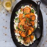Charred sweet potato rounds and herbs served on a bed of whipped goat cheese, sprinkled with dukkah and fried shallots, presented on a black oval platter with a rustic black spoon and small glasses of amber liquid.