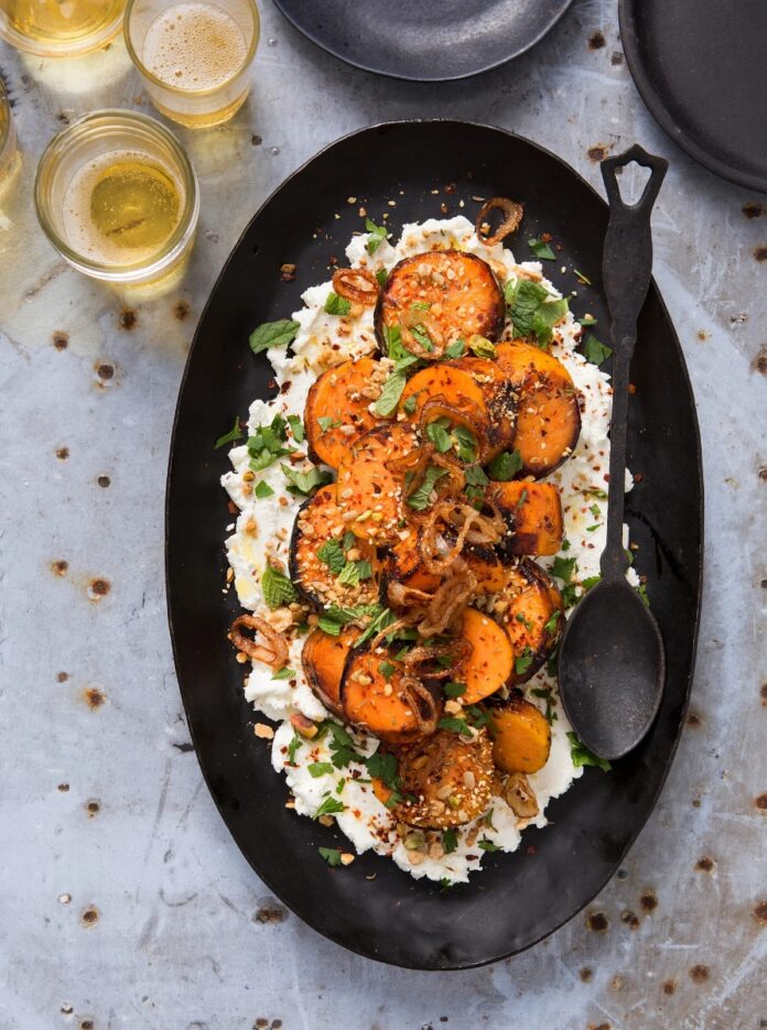 Charred sweet potato rounds and herbs served on a bed of whipped goat cheese, sprinkled with dukkah and fried shallots, presented on a black oval platter with a rustic black spoon and small glasses of amber liquid.