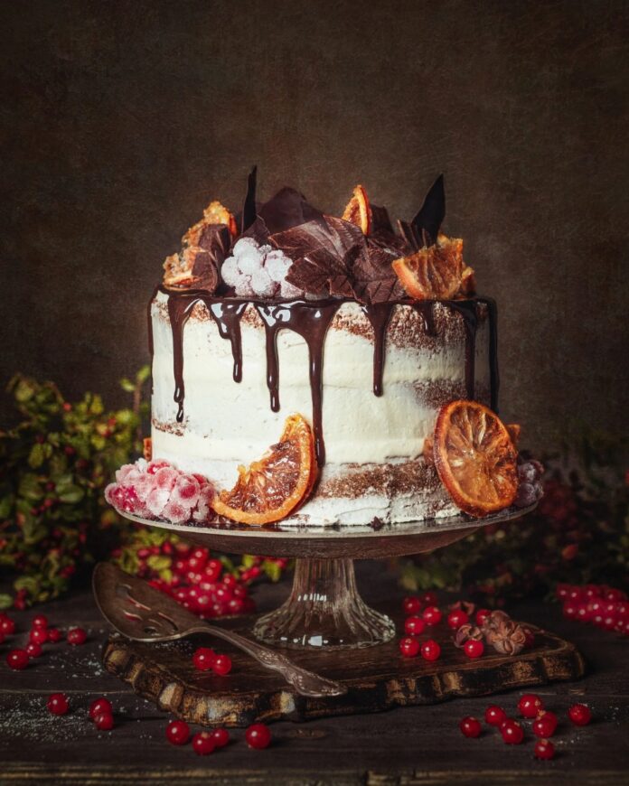 A stunning three-layer Chocolate Sponge Cake on a pedestal, iced with white buttercream and chocolate ganache drip, decorated with candied orange slices, chocolate shards, and frosted red currants for a festive winter dessert.