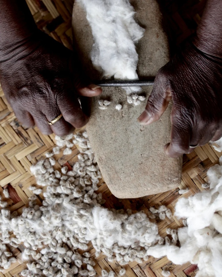 Dark-skinned hands using a round metal bar and a grindstone to manually tease and separate cotton seeds from white cotton fibers on a woven mat, part of the traditional Malian mudcloth process.