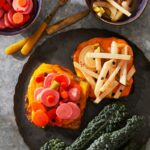 A dark plate with toast topped with bright pink and orange escabeche (pickled carrots and radishes) and dark green kale, with two small bowls on the side.