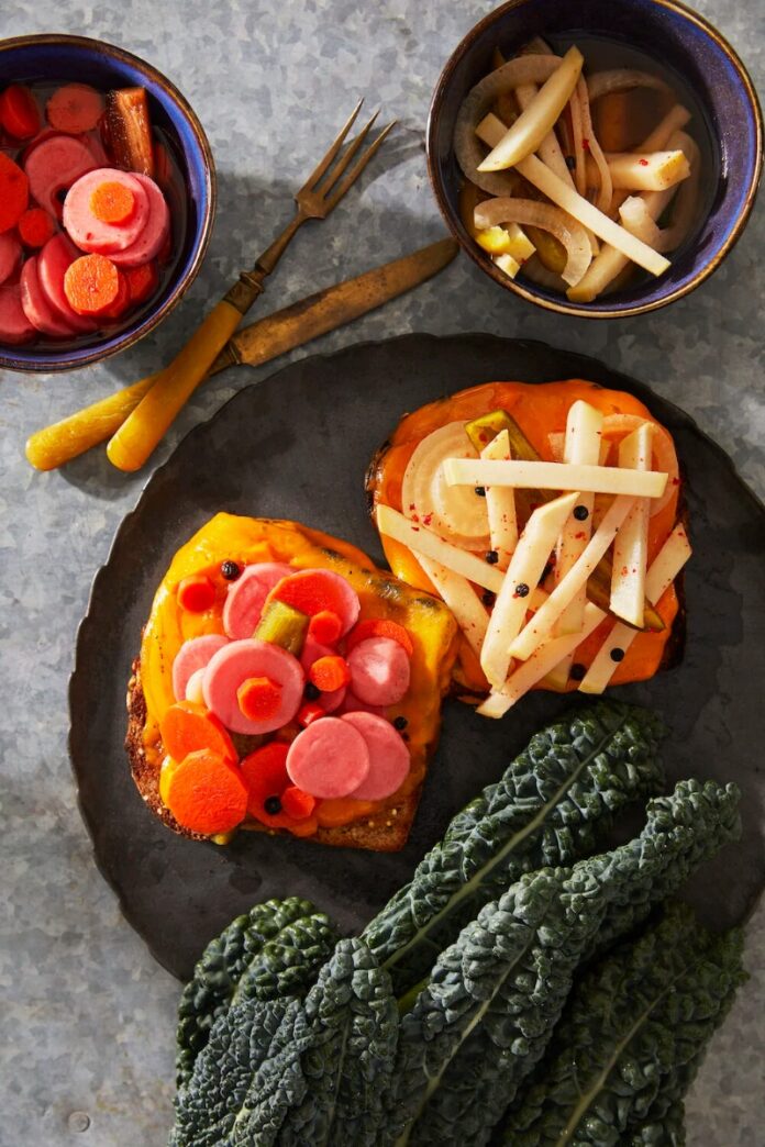 A dark plate with toast topped with bright pink and orange escabeche (pickled carrots and radishes) and dark green kale, with two small bowls on the side.