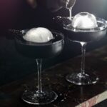 Two martini glasses with two sphere ice balls as a person pours something into the back one.