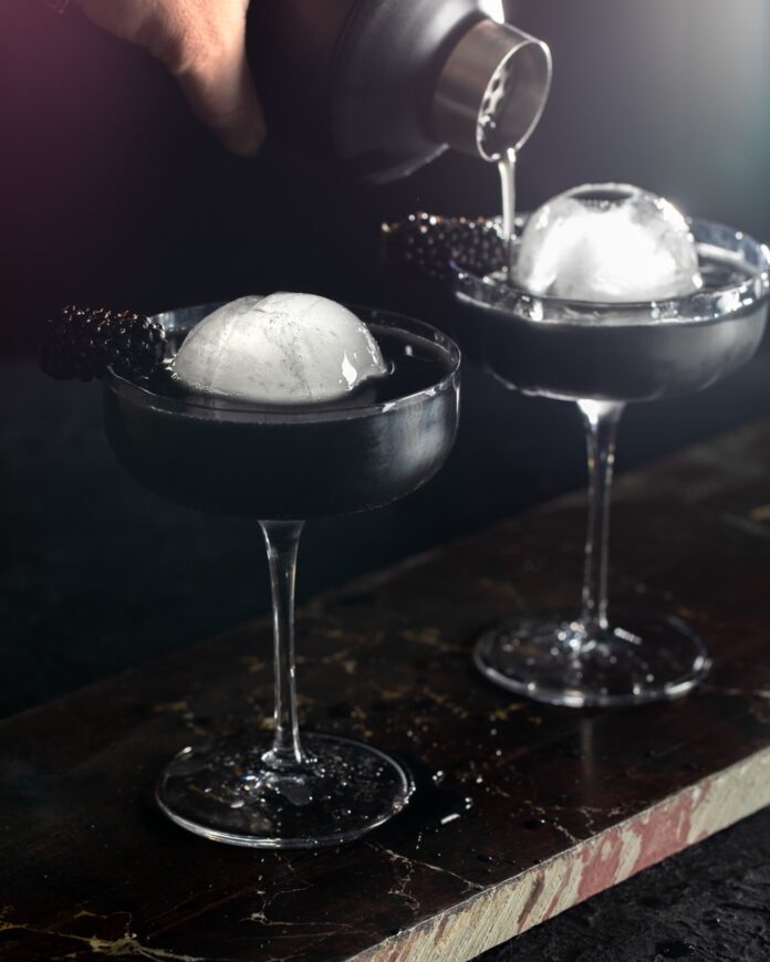 Two martini glasses with two sphere ice balls as a person pours something into the back one.