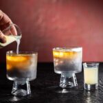 Someone pours a yellow liquid into a dark cocktail.