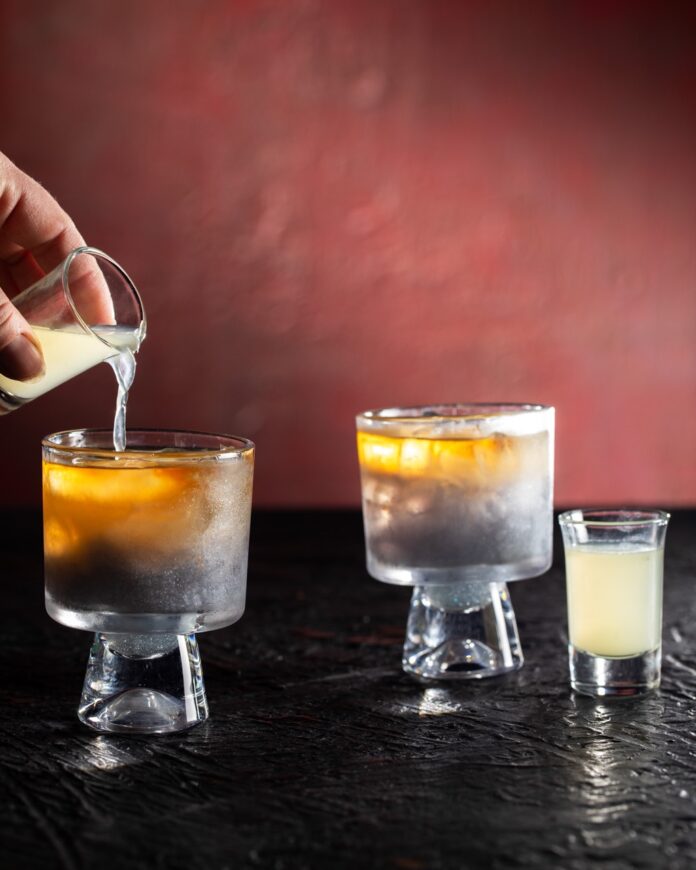 Someone pours a yellow liquid into a dark cocktail.