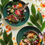 Two deep-green Villeroy & Boch plates holding fried pickled mackerel garnished with seared bok choy, radish, and colorful fresh vegetables, surrounded by bright green leaves and flower petals.