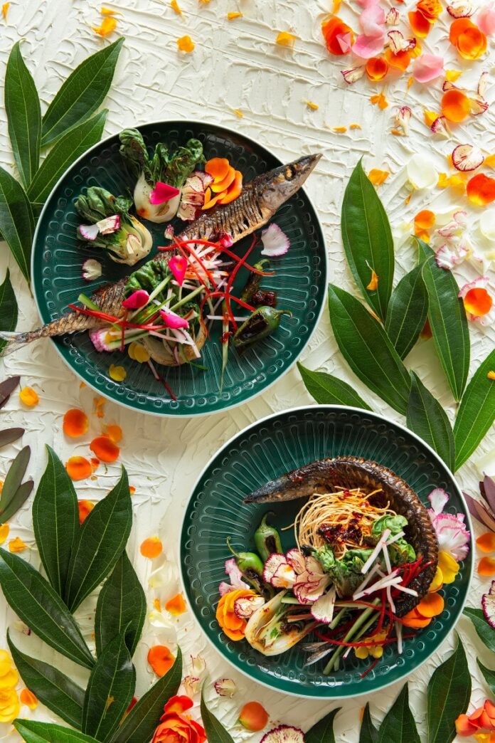 Two deep-green Villeroy & Boch plates holding fried pickled mackerel garnished with seared bok choy, radish, and colorful fresh vegetables, surrounded by bright green leaves and flower petals.