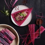 A small white plate with a hot pink tamale in a triangle shape and rolled tamales on a plate in the bottom left corner.