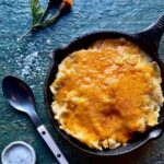A cast iron pan of Green Chile and Cheddar Mashed Potatoes with a spoon, salt, and orange flowers beside the pan.