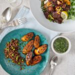 A haitian food featuring a turquoise plate with grilled plantains and pigeon peas and rice (diri ak pwa), alongside a small white bowl of bright green Epis seasoning base, and a white plate with fried pork (griot) and slaw.