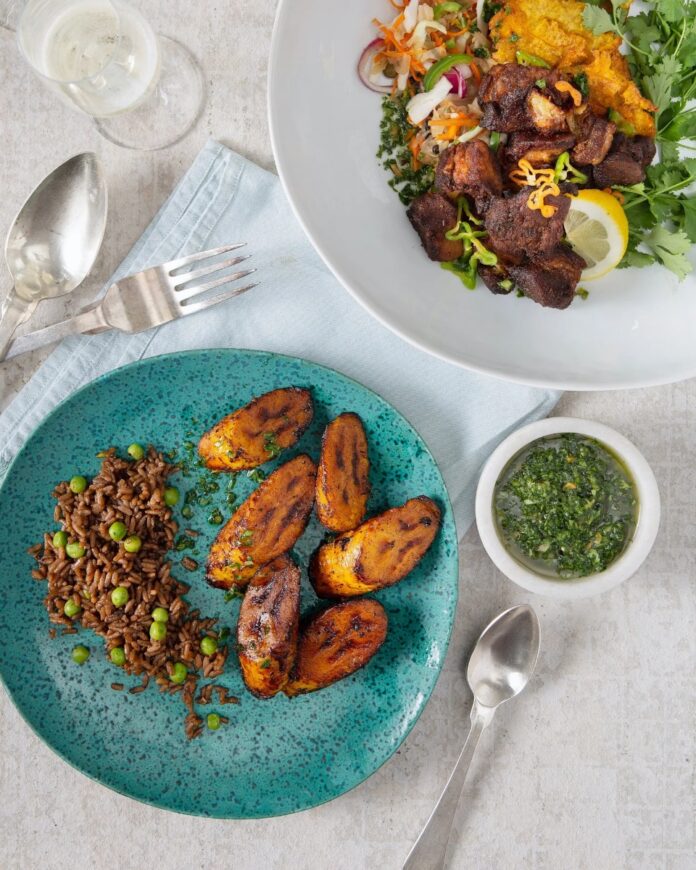 Haitian Epis TABLE Magazine A haitian food featuring a turquoise plate with grilled plantains and pigeon peas and rice (diri ak pwa), alongside a small white bowl of bright green Epis seasoning base, and a white plate with fried pork (griot) and slaw.