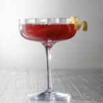 A cocktail in a martini glass in red color with a lemon twist garnish.