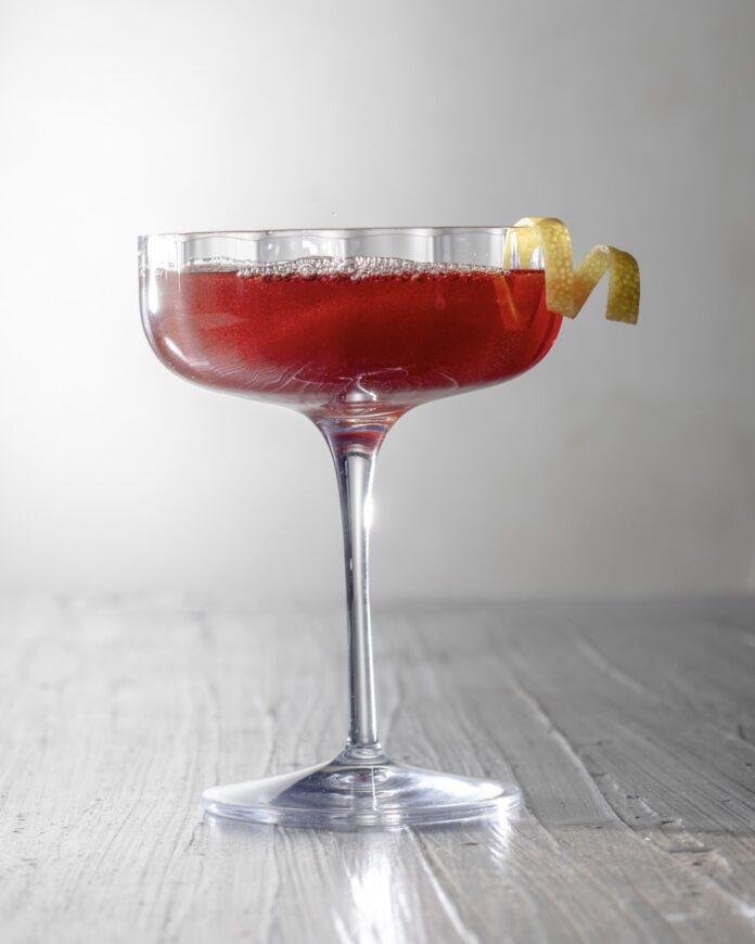 A cocktail in a martini glass in red color with a lemon twist garnish.