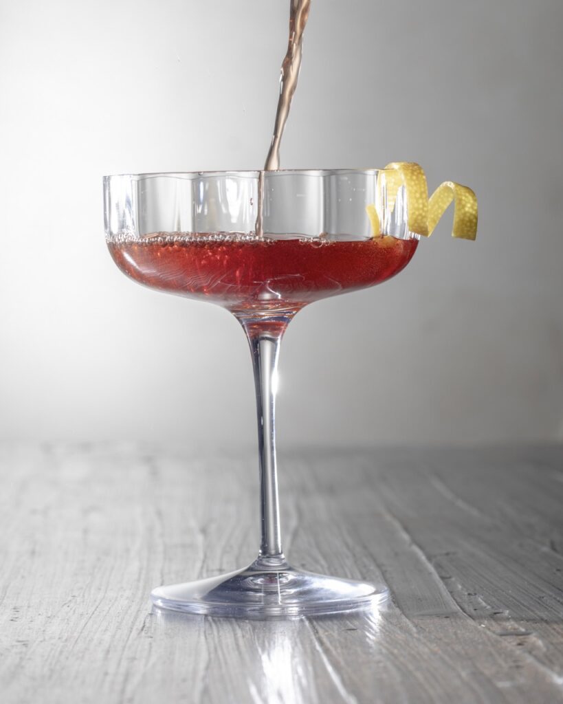 Red kosher cocktail being poured into a martini glass with a lemon twist.