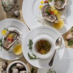 A flat lay photo of fresh oysters on the half shell served on white Wedgwood plates with salt and mignonette sauce, garnished with chili peppers, scallions, and lemon slices.