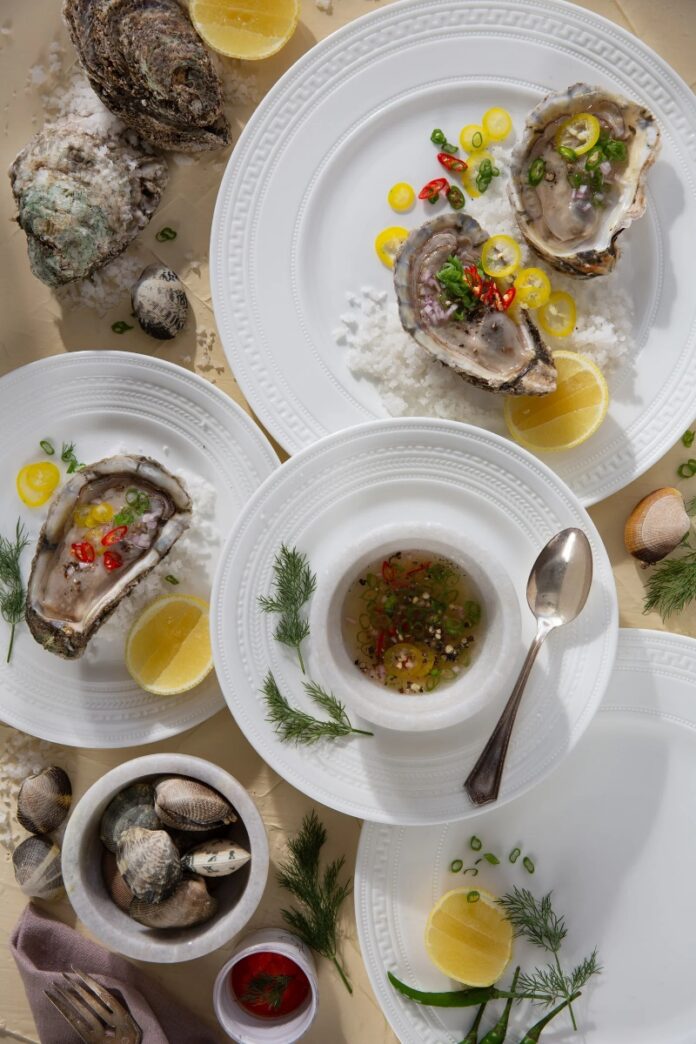 Mignonette TABLE Magazine A flat lay photo of fresh oysters on the half shell served on white Wedgwood plates with salt and mignonette sauce, garnished with chili peppers, scallions, and lemon slices.