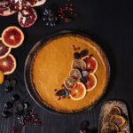 A miso sweet potato pie sits on a dark wood table with sliced citrus, grapes, and pomegranate seeds on top.