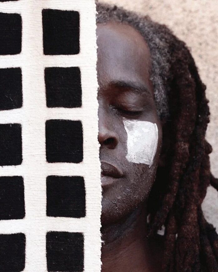 A man with dark dreadlocks and white clay paint on his cheek, partially obscured by a piece of black and white checkered bògòlanfini (Malian mudcloth), showcasing the traditional textile craft.