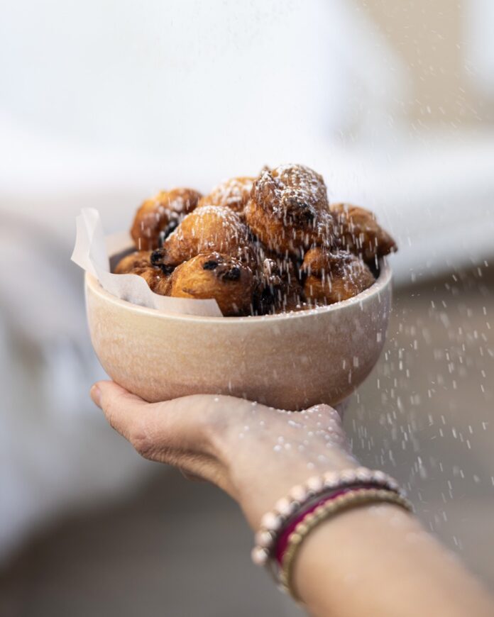 A white bowl full of Oliebollen Dutch donuts being held by a person as another sprinkles powdered sugar overtop.