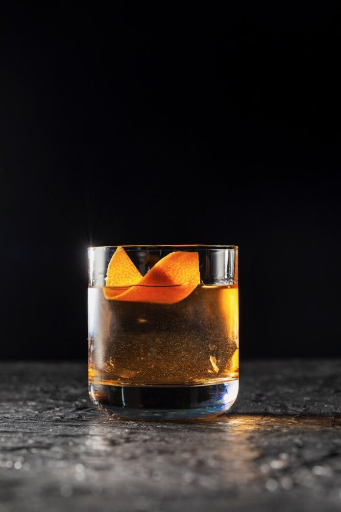 A short glass with a clear old fashioned for the release of Now You See Me Now You Don't with an orange peel twist as a garnish.