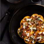 A persimmon, burrata, and fried shallot salad on a large black plate, garnished with basil and a small black fork and knife, with a glass of dark iced beverage.