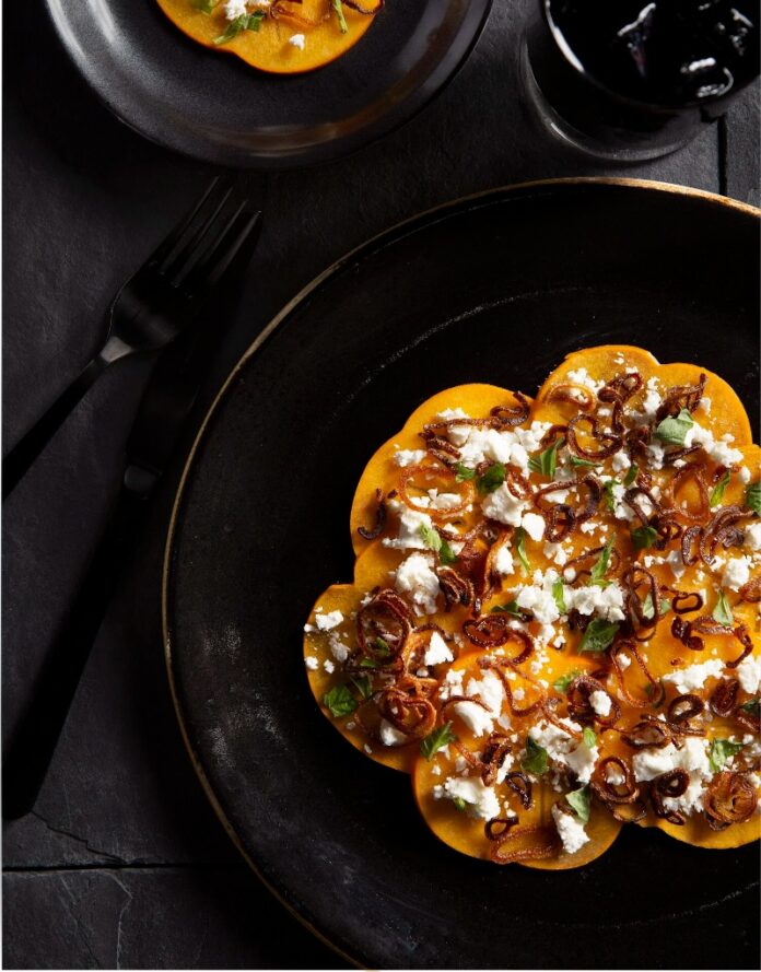 Persimmon, Yuzu, Basil, Fried Shallot, & Burrata TABLE Magazine A persimmon, burrata, and fried shallot salad on a large black plate, garnished with basil and a small black fork and knife, with a glass of dark iced beverage.